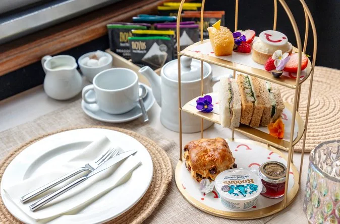 A tiered stand of scones, sandwiches and cakes beside a teapot on a set table